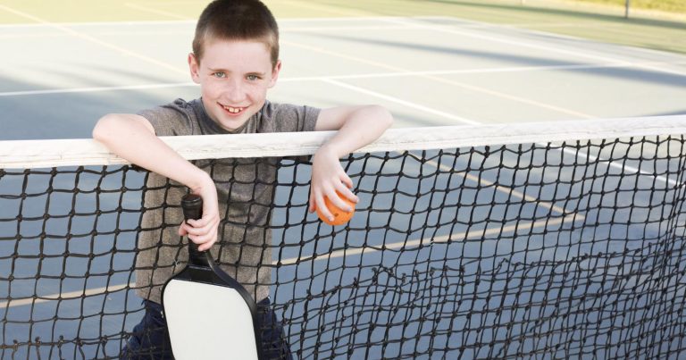Pickleball For Kids: 7 Reasons It’s The Most Kid-Friendly Sport Young boy smiling holding a pickleball paddle and leaning against a pickleball net